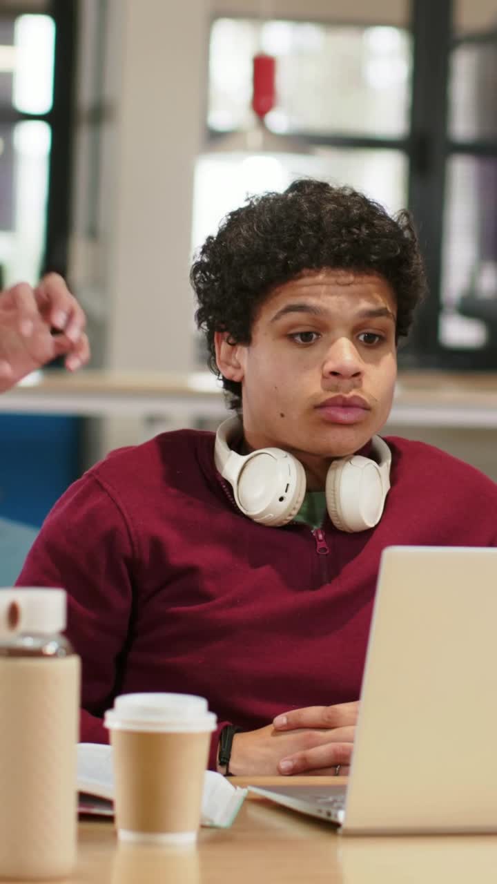 Vertical video: Turning man in maroon sweater listening to waving hand at study table with laptop