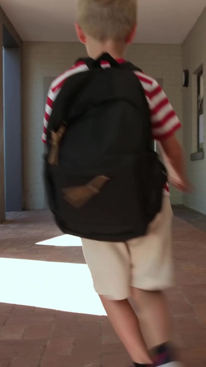Walking boy in striped tee carrying black backpack spotting sunlit courtyard, quickening into skip