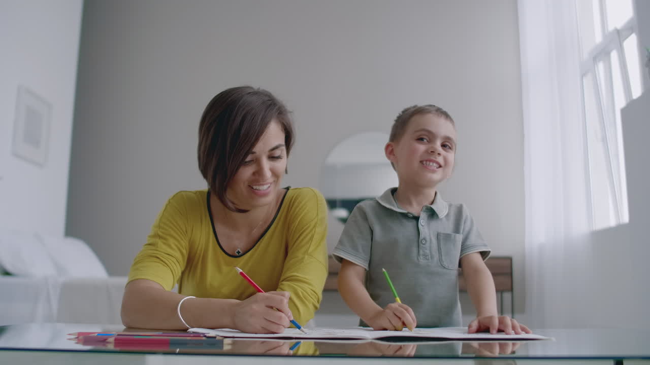 Mom and son sitting at the mirror table in the white room draw with colored pencils smiling and laughing