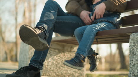 close-up of an adult and a child sitting closely together on a bench, the child playfully dangles his legs, gently hitting the adult s legs, the blurred background of trees and an electric pole