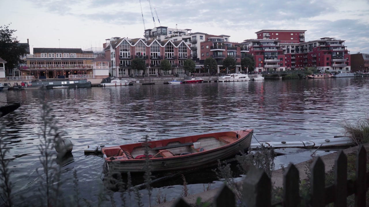 A small wooden boat rests on calm water near a picket fence, with riverside apartments and waterfront buildings across the channel under a muted sky.