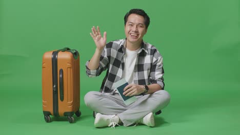 full body of asian male traveler with luggage and passport smiling, waving hand, and saying bye while sitting in the green screen background studio