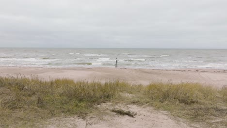 establishing aerial view of baltic sea coast on a overcast day, frame jurmalciems, old wooden pier, white sand beach, large storm waves crushing against the coast, wide drone shot moving backward