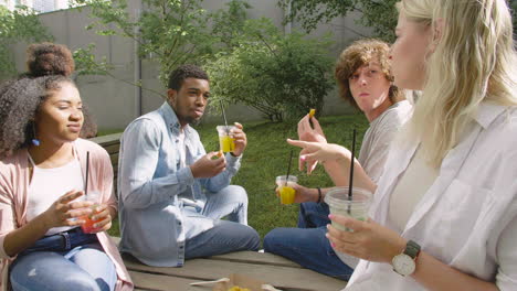 Young-Blonde-Woman-Talking-With-Her-Multiethnic-Friends,-Sitting-In-A-Wooden-Bench-In-A-Park,-Drinking-And-Eating-Together