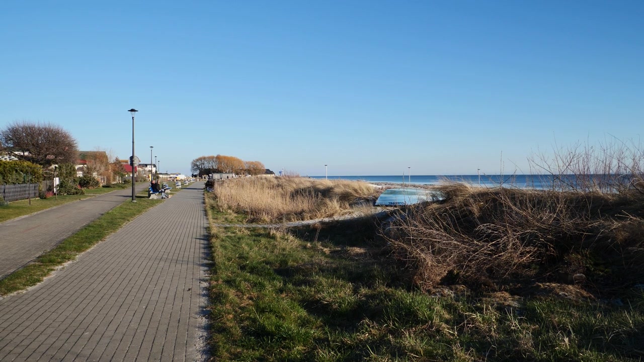 Eine leere Strandpromenade in Kuźnica, Polen, leuchtet unter einem warmen Sonnenuntergang. Ruhige Ostseewellen, Küstengras und friedliche, leere Bänke fangen die besinnliche Nebensaison-Einsamkeit ein.