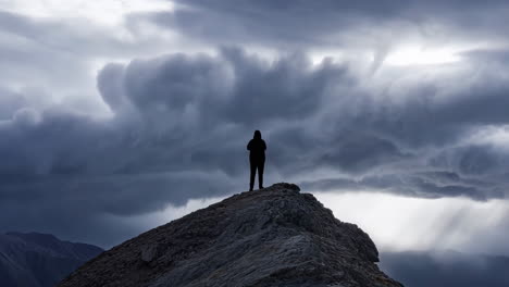 a solitary figure stands atop a mountain peak under a dramatic, stormy sky