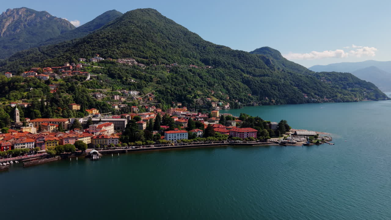 Drone performs a panning arc along the curved lakeside, showing colorful buildings, trees, and surrounding mountains on Lake Como in Italy