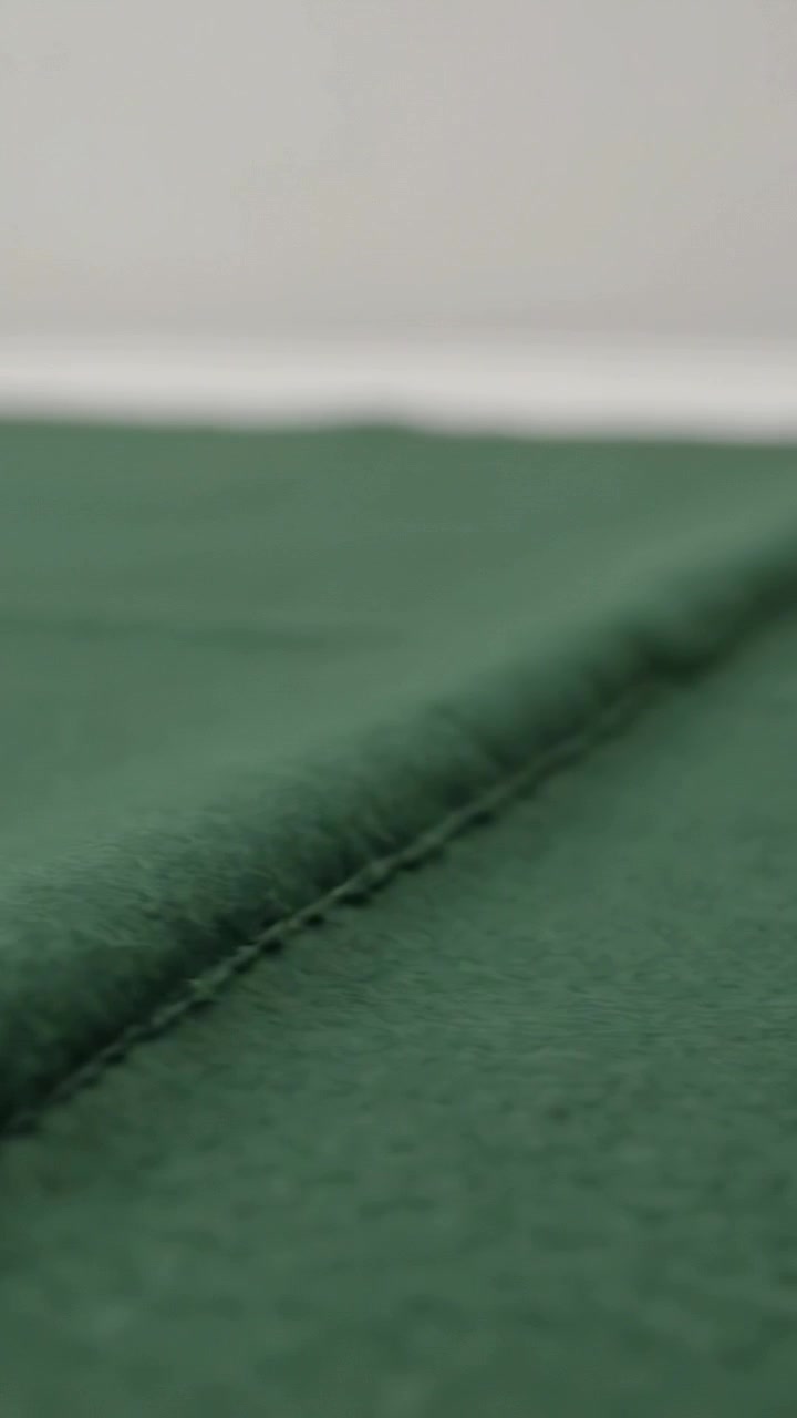 Vídeo vertical: Câmera focando em uma costura de tecido verde em uma mesa contra uma parede clara, destacando a costura.