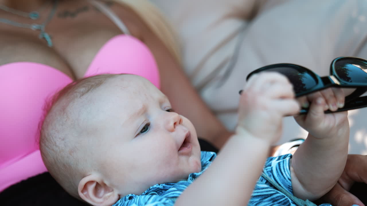 Lovely Caucasian infant lies on his mom outdoors. Adorable baby is playing with mom's sunglasses. Close up.