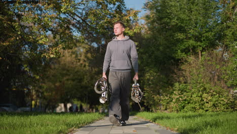 young man with short brown hair walking on paved path holding spring-loaded stilts, wearing light gray hoodie and olive-green cargo pants, surrounded by green trees and grass in bright natural setting