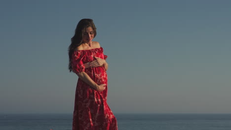 disparo medio de una mujer embarazada con un vestido rojo sosteniendo su vientre cerca del mar