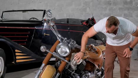 man polishing a vintage motorcycle next to a classic car in a garage