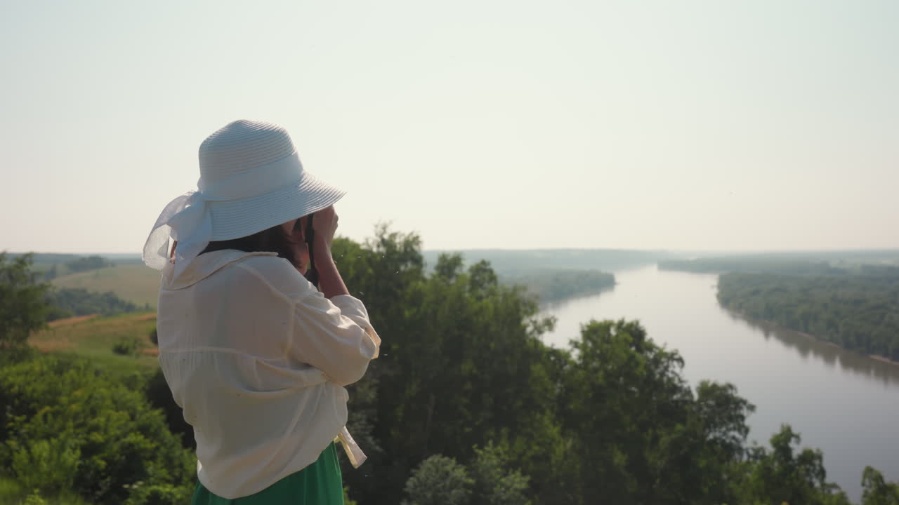 Donna con cappello bianco si trova su un punto panoramico elevato, fotografando un fiume suggestivo che serpeggia attraverso una lussureggiante foresta verde sotto un cielo diurno velato.