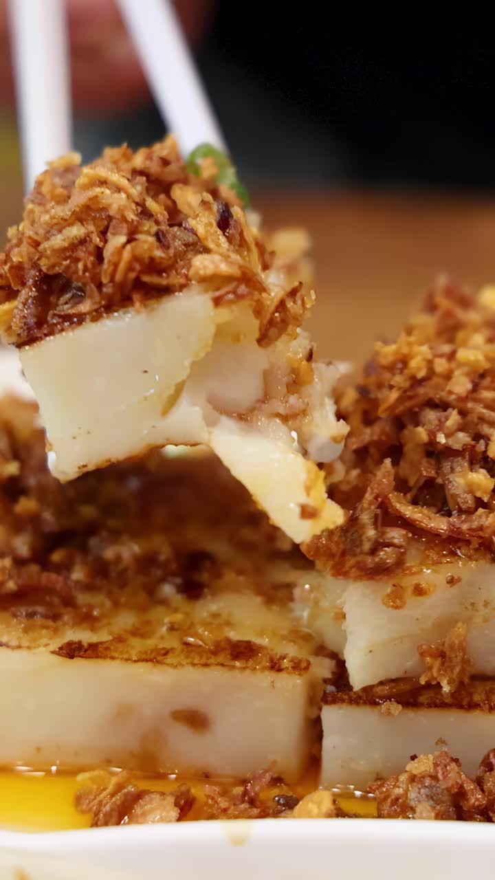 Delle bacchette afferrano un pezzo di torta di rapa saltata in padella, guarnita con scalogni fritti croccanti, da un piatto in un ambiente da pranzo illuminato calorosamente. Primo piano, profondità di campo ridotta.
