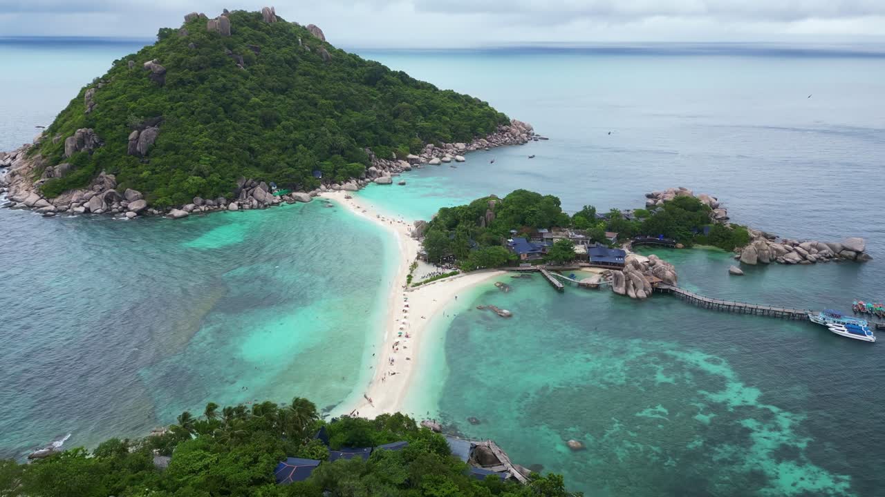 Aerial view of a tropical beach with clear turquoise waters, sandy shores, lush green hills, and a rocky coastline.