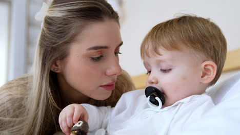 mother talks to her little baby boy sucking a pacifier. cute toddler is holding a toy and looks at it.
