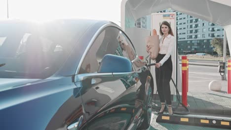 hermosa mujer de negocios se acerca a su coche eléctrico con comestibles y mira a la cámara, sonriendo. carga de coche eléctrico. transporte respetuoso con el medio ambiente. carga automática finalización. compras