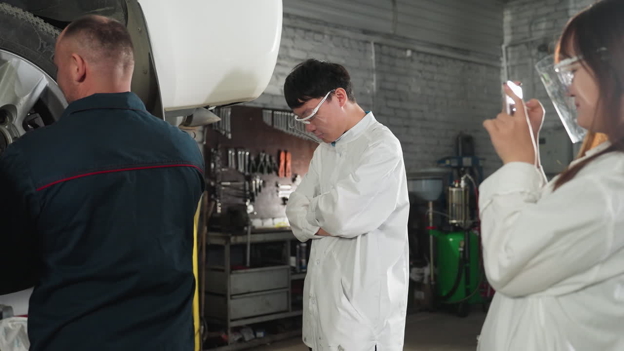 inventor en bata de laboratorio tomando una foto de un mecánico arreglando un neumático de coche mientras el estudiante observa con los brazos cruzados en el taller de mecánicos, herramientas en el fondo
