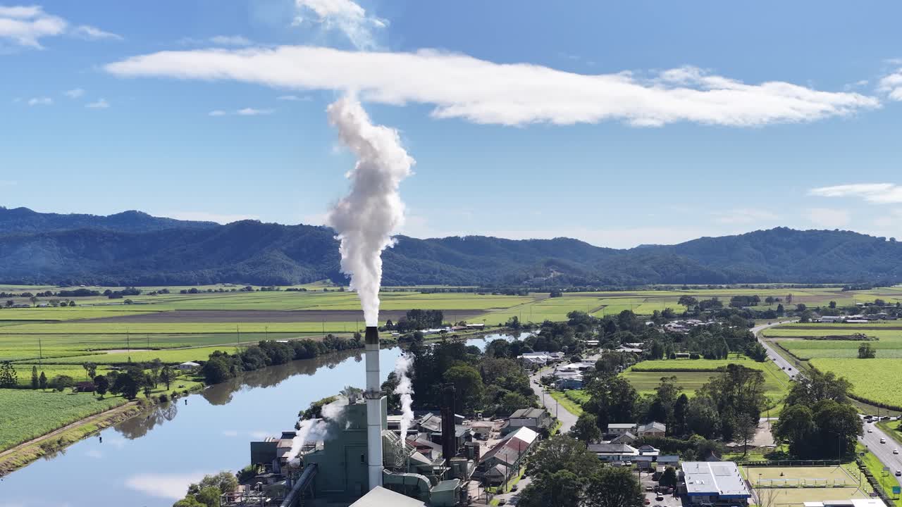 Aerial footage of a sugar mill with smoke rising, set against a lush, rural landscape under clear skies