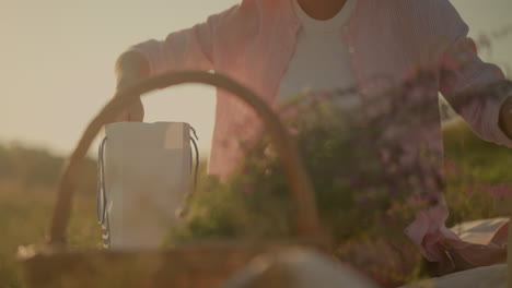 lady seated on picnic mat outdoors placing fresh croissant on plates atop wooden picnic table, nearby, a basket filled with flowers adds to the scenic sunlit background