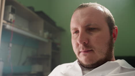 a head view of a technician in a white lab coat, looking thoughtful and focused in a laboratory setting, with a shelf fill with some equipment