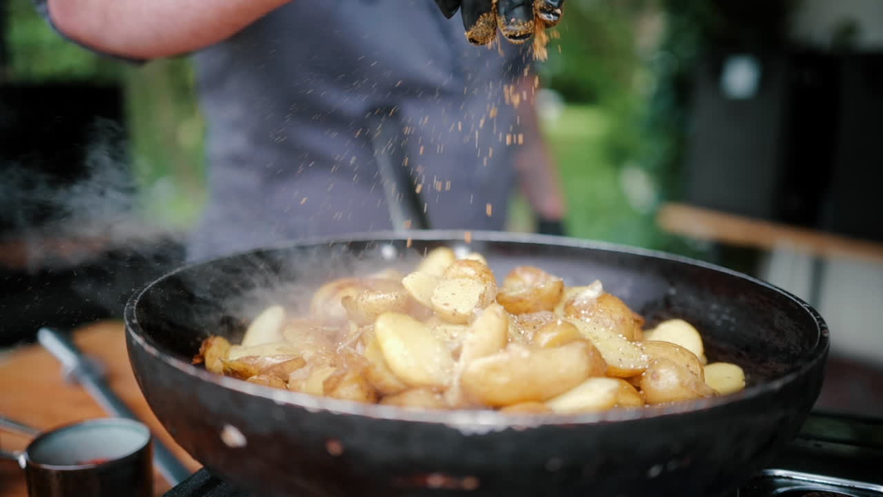Koks hånd krydrer kartofler på en pande på et gasblus ved udendørs catering til et arrangement, i slowmotion nærbillede.