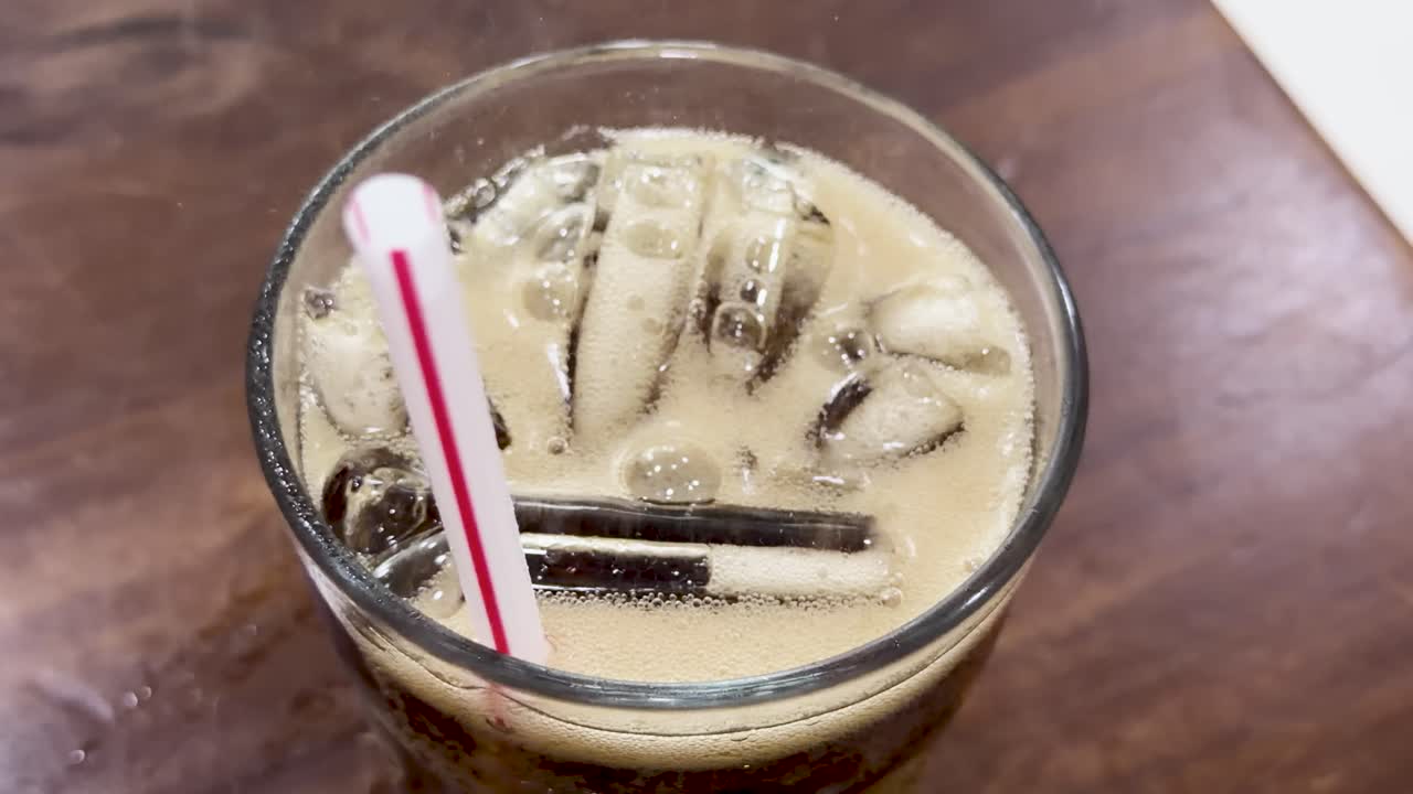 Dark fizzy cola pours from a red can into a glass with ice and straw