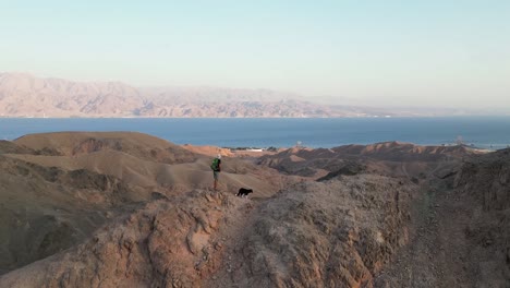 una vista de un hombre caminando con su perro en las montañas del desierto, con una vista al mar capturada por un dron