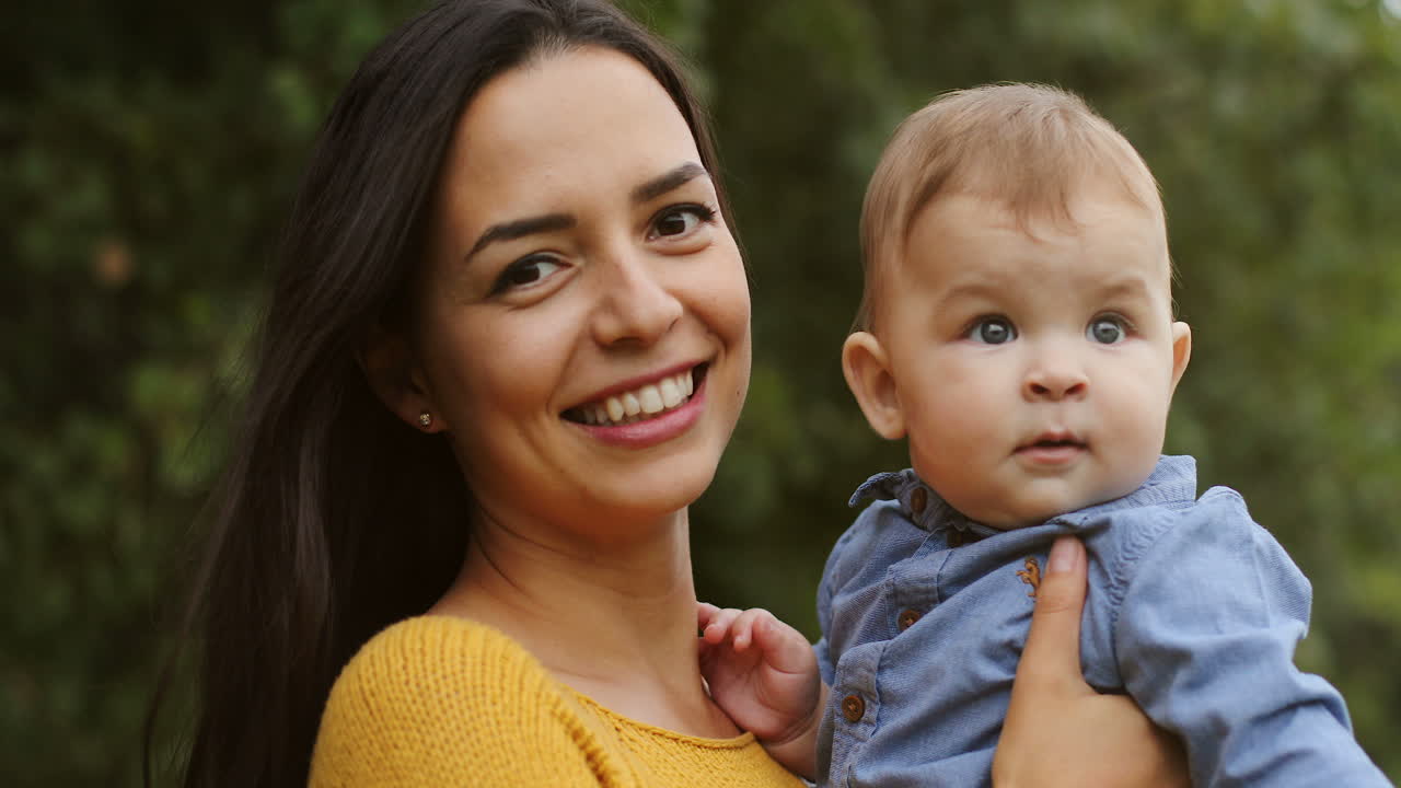 Portrætfoto af en smilende ung kvinde med langt mørkt hår, der holder sin søde lille dreng og kigger ind i kameraet i parken.