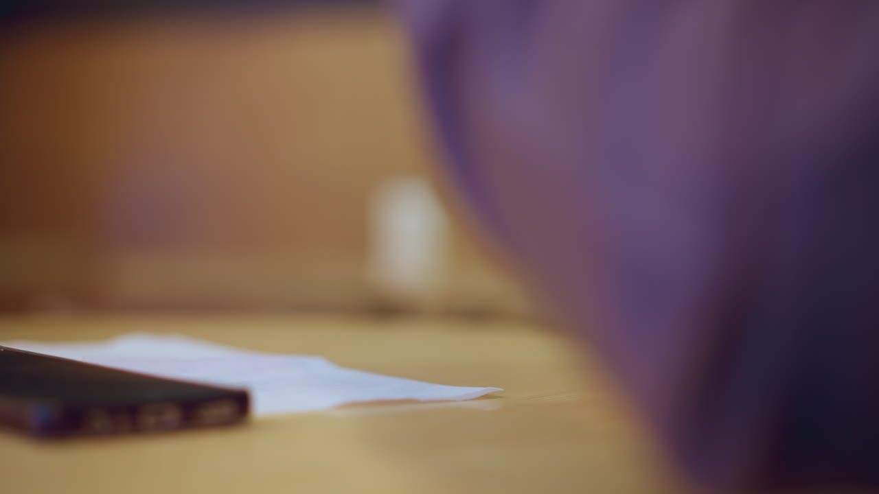 Close-up view of smartphone resting on napkin atop wooden table with blurred arm in purple shirt in foreground, creating intimate indoor atmosphere with soft lighting and subtle details in casual setting