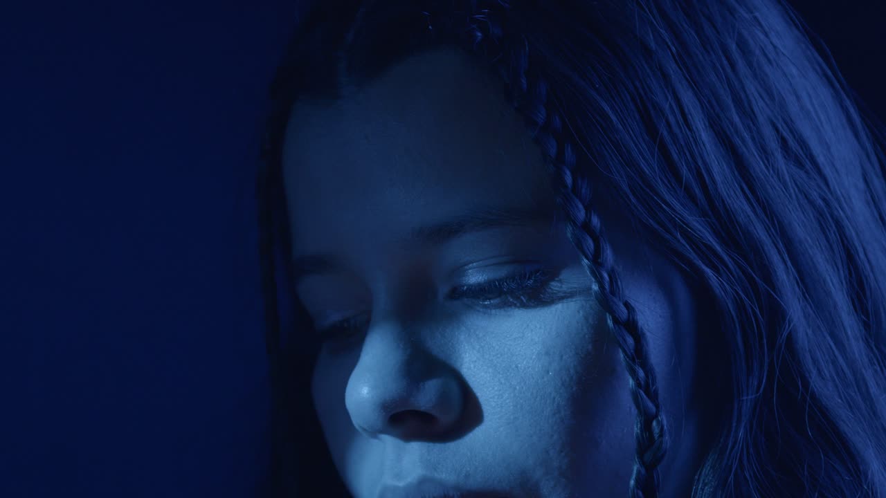 Close-up shot of a contemplative woman under moody, deep blue lighting. The braided hair and solemn expression convey a sense of introspection and emotion