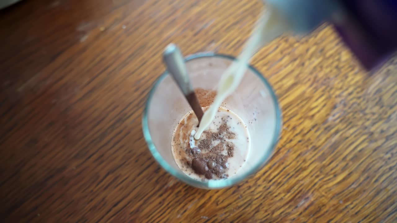 Primo piano dall'alto di latte versato in un bicchiere con cacao in polvere e un cucchiaio su un tavolo di legno, preparazione di una bevanda al cioccolato fatta in casa