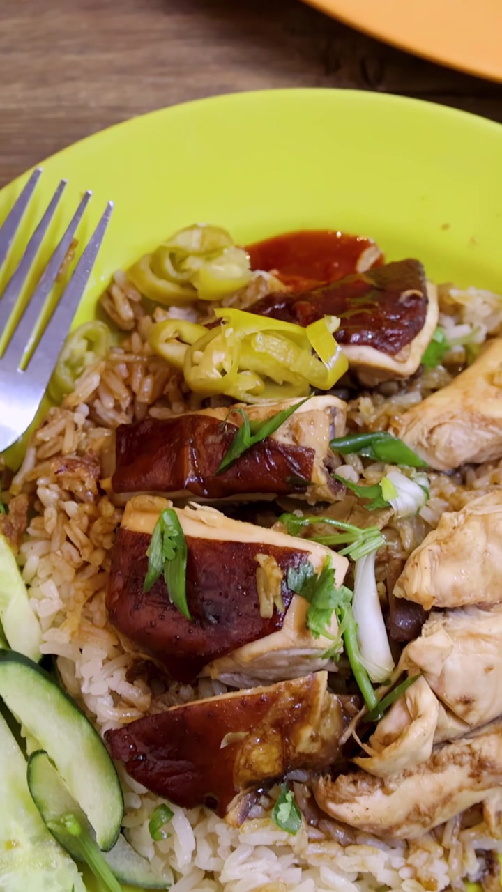 La telecamera si muove fluidamente da una ciotola di zuppa di noodle arrostiti a un piatto di riso al pollo con verdure su un tavolo di legno sotto un'illuminazione brillante e uniforme