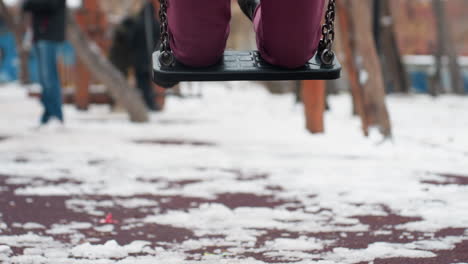 close-up of person legs kneeling on iron swing, wearing black boots in snowy outdoor park, blurred background features other people playing and swinging