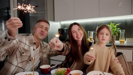 Portrait-of-a-happy-brunette-woman-in-a-plaid-shirt-together-with-her-husband-and-little-daughter-with-a-braided-hairstyle-who-are-holding-sparklers-in-their-hands-having-fun-and-rejoicing-while-sitting-at-the-dining-table-during-a-festive-dinner-in-the-evening-in-a-modern-apartment