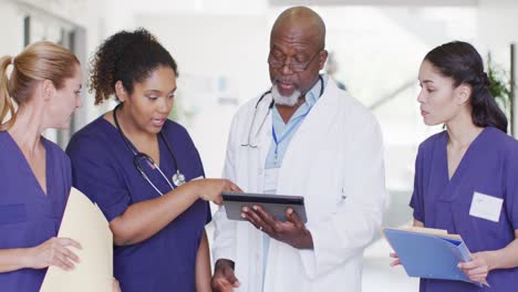 Video-of-diverse-male-and-female-doctors-looking-at-tablet-and-files,-talking-in-hospital-corridor