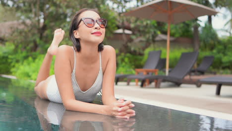 lying on her stomach and kicking her feet slowly in the air, an attractive young woman in sunglasses enjoys the amenities a resort has to offer