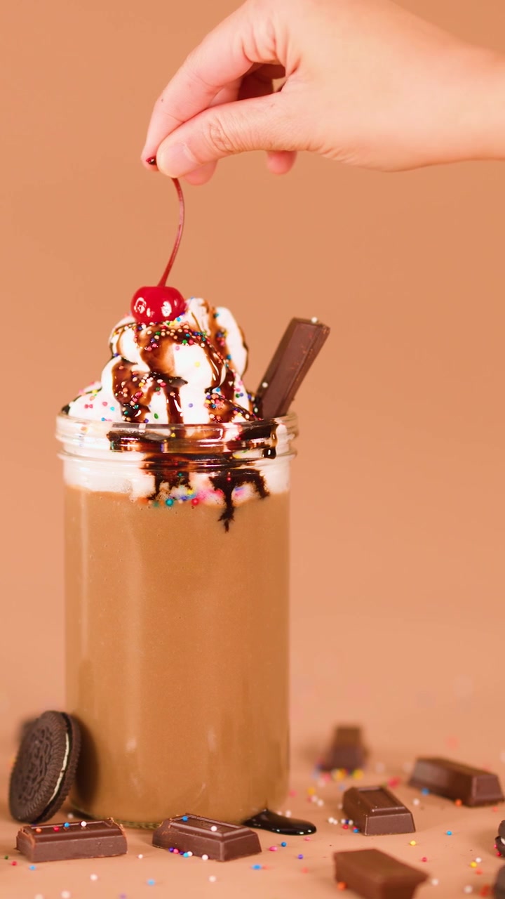 Una mano posiziona una ciliegia sulla cima di un milkshake al cioccolato con panna montata, sciroppo al cioccolato, biscotti e codette di zucchero