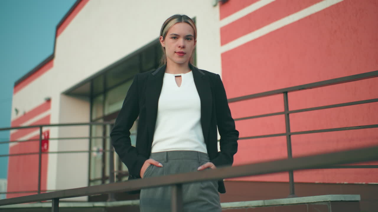 Donna sicura di sé in blazer nero e top bianco posa con le mani in tasca vicino a una ringhiera di ferro di fronte a un moderno edificio per uffici, trasmettendo eleganza, professionalità e compostezza sotto un cielo sereno