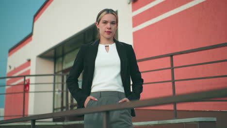 confident lady in black blazer and white top poses with hands in pockets near iron railing in front of modern office building, exuding elegance, professionalism, and poise under clear sky