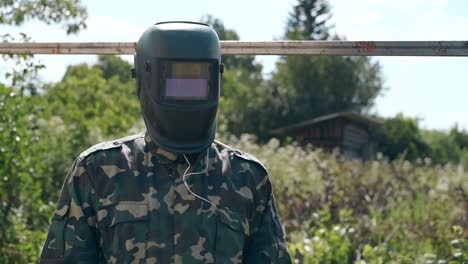 worker in camouflage takes off welding mask at fence