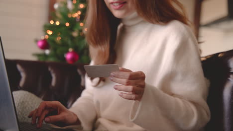 Close-Up-View-Of-Woman-Hands-Who-Is-Shopping-Online-Using-Credit-Card-While-Sitting-On-The-Sofa-Near-A-Christmas-Tree-In-The-Living-Room-At-Home