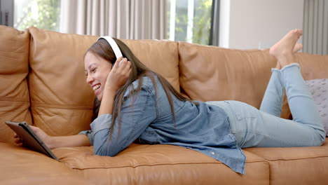 biracial young woman wearing headphones, lying on couch, holding tablet