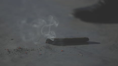 close up shot of dropping a cigar in slowmotion on a concrete floor with burning sparks coming off and smoke rising in the air while a person and shoe is walking away in the background log