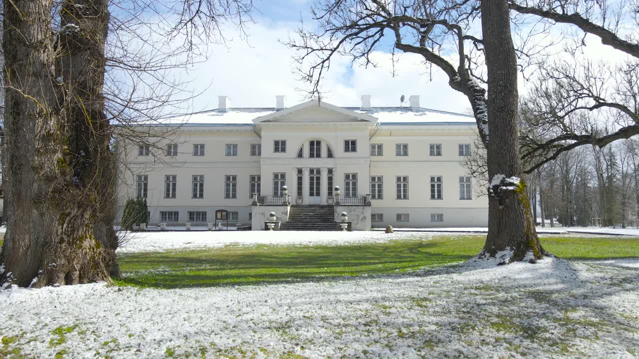 Gorgeous footage slowly approaching and moving closer to Saku mansion that has big pillars and the building is white. Grass is green and some white snow is glittering in the sunny day, blue sky also.
