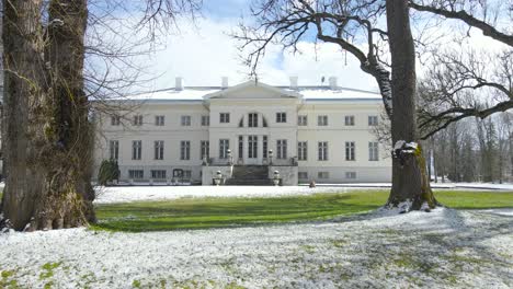 gorgeous footage slowly approaching and moving closer to saku mansion that has big pillars and the building is white. grass is green and some white snow is glittering in the sunny day, blue sky also.
