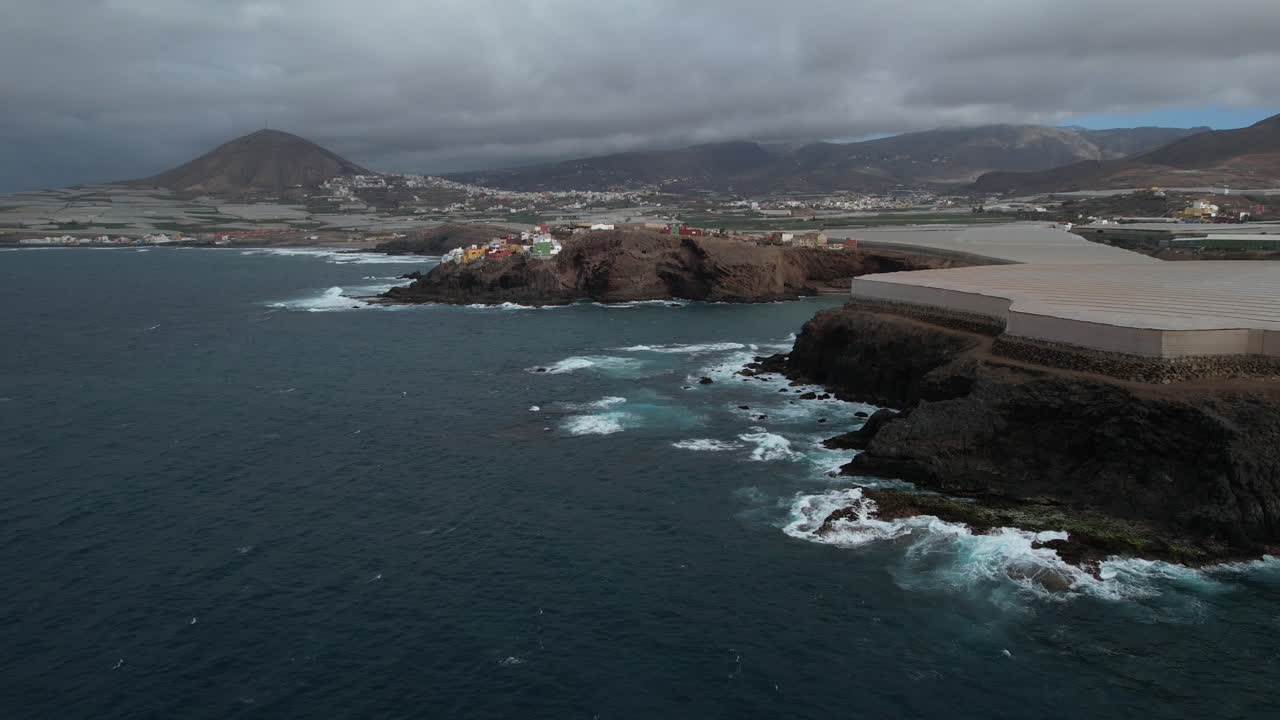 maravillosa toma aerea en orbita en el lugar conocido como punta de galdar y sobre invernaderos, con el monte galdar al fondo y las casas construidas en la costa