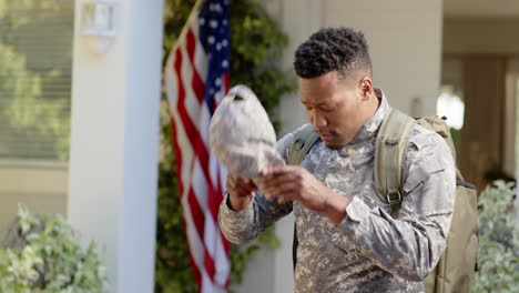 serious african american male soldier putting cap on over flag of usa at home, slow motion