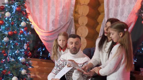 father and mother with two daughters in an armchair near a christmas tree with decorations are considering children's drawings. new year's celebration