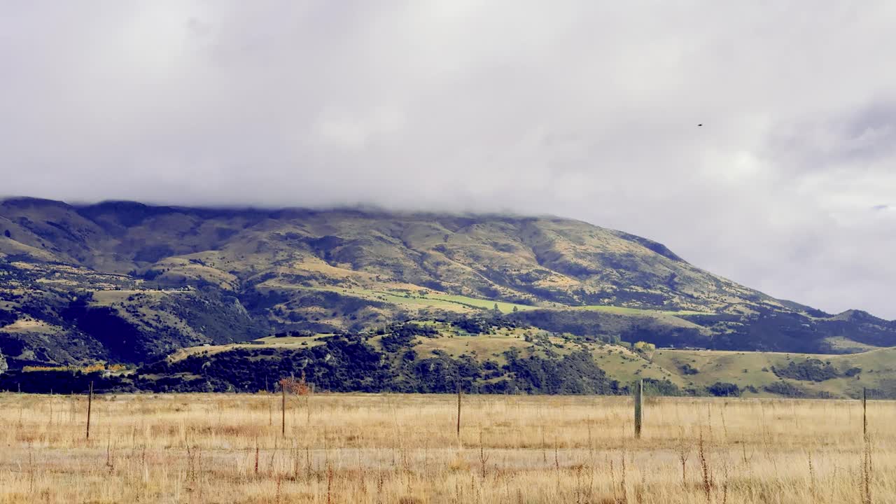 A moving vehicle captures golden grass fields and rugged mountains under cloudy skies, with smooth lateral camera motion and soft, natural daylight
